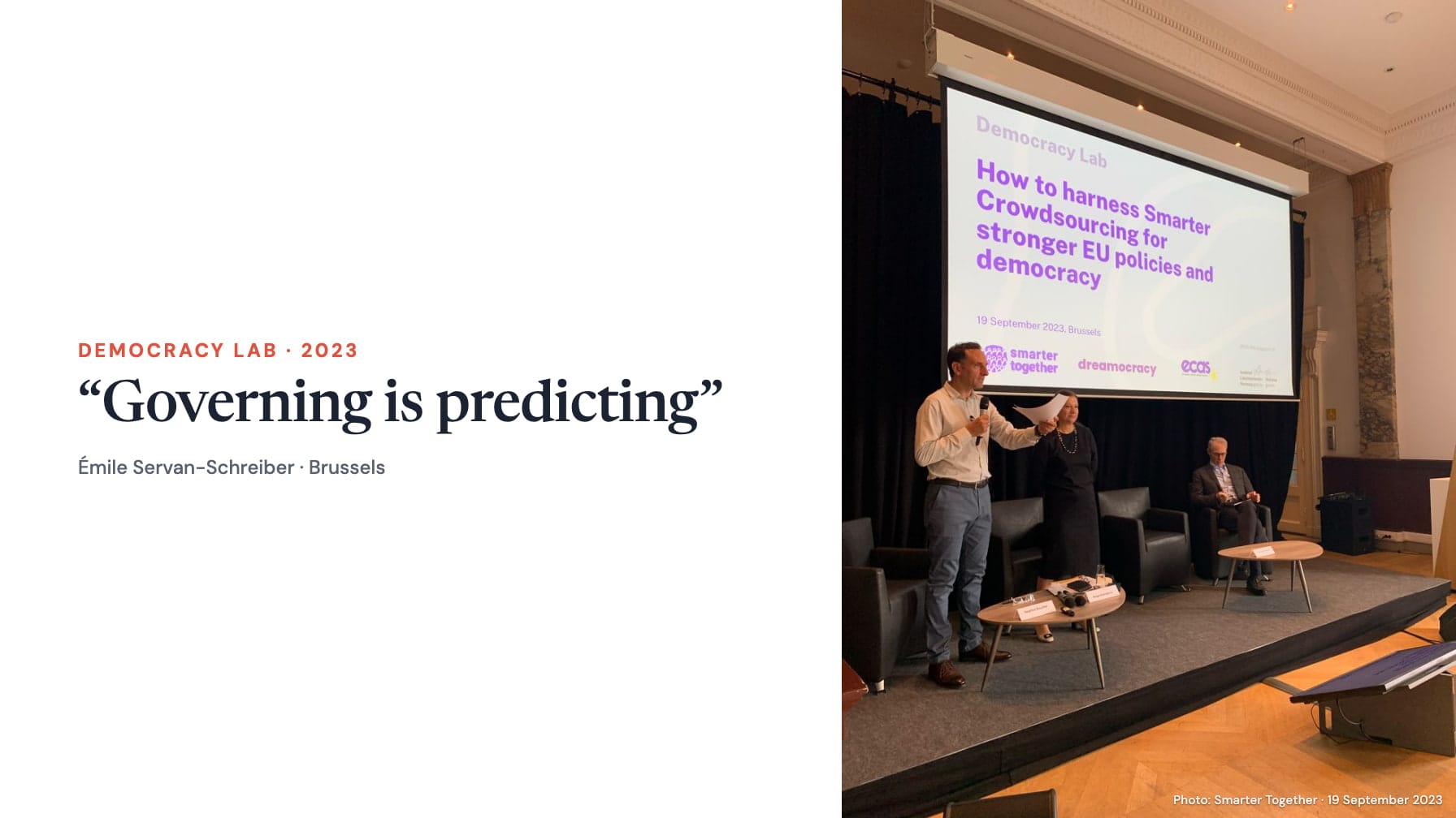 A white panel reading “Democracy Lab · 2023” and “Governing is predicting”, attributed to Émile Servan-Schreiber in Brussels, beside the complete event photograph of three people on a stage beneath the Democracy Lab screen; photo credited to Smarter Together, 19 September 2023.