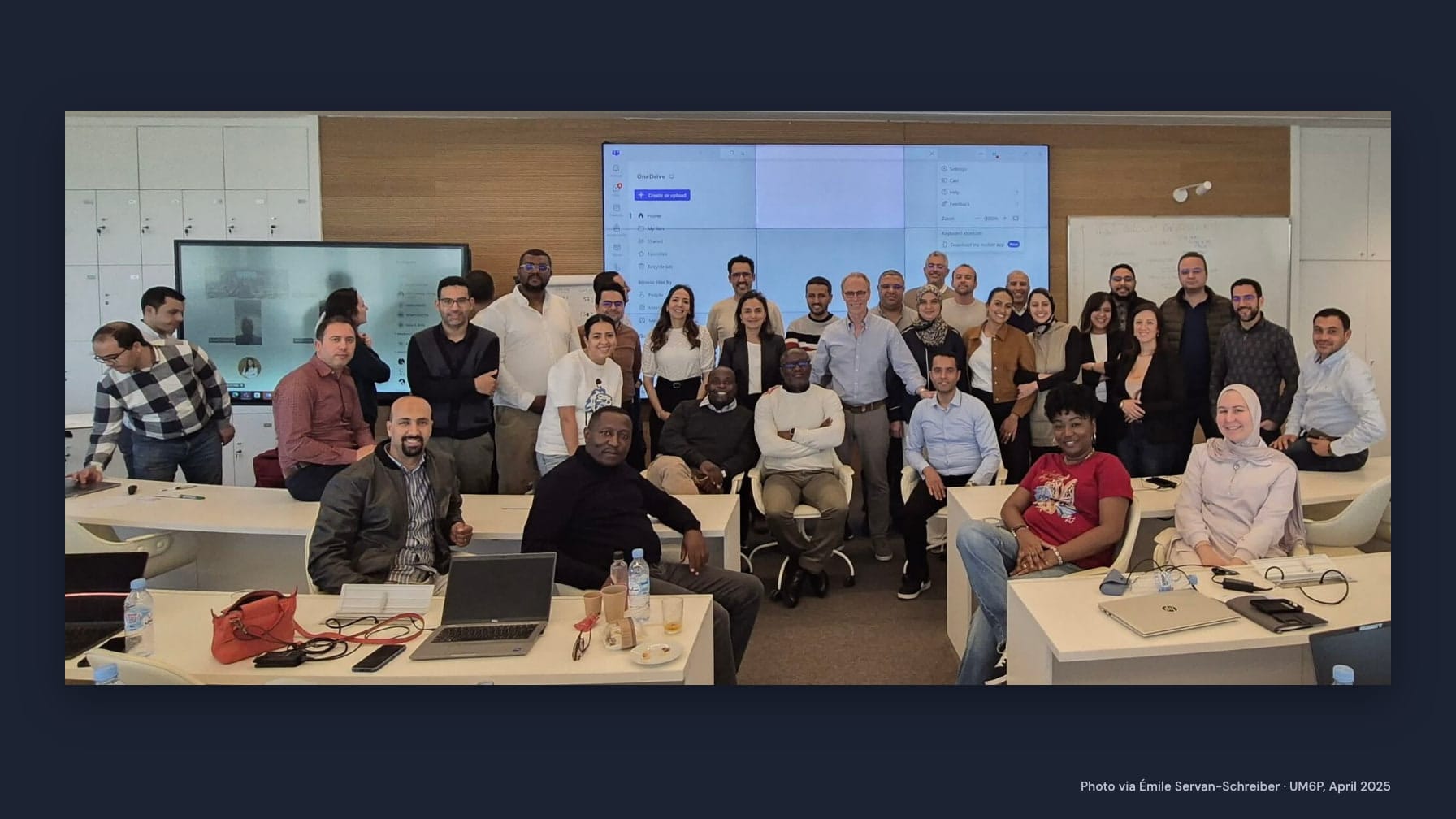 Émile Servan-Schreiber, standing in a light blue shirt, and Executive MBA students pose together in a classroom with desks, laptops and a large screen, framed by navy with the credit “Photo via Émile Servan-Schreiber · UM6P, April 2025”.